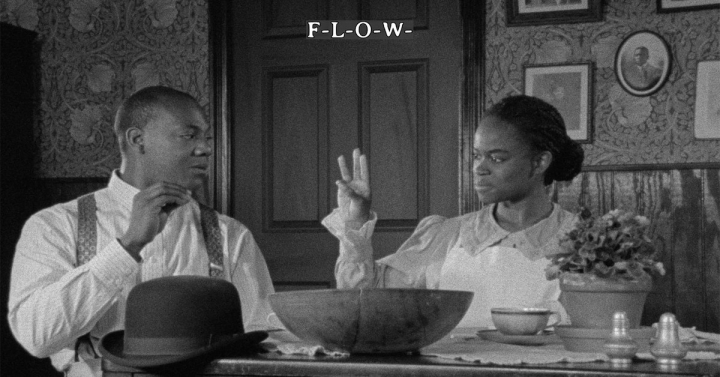 Black and white image of woman teaching man sign language, holding up her hand to demonstrate the word 