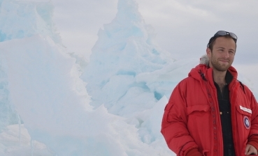 David Lesser stands against an icy Antarctic backdrop wearing a heavy red jacket. 