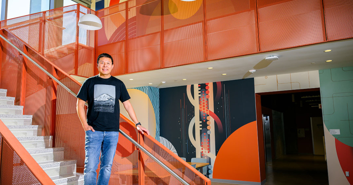 A man, Brian Choi, stands on a staircase in front of a colorful wall mural he created.