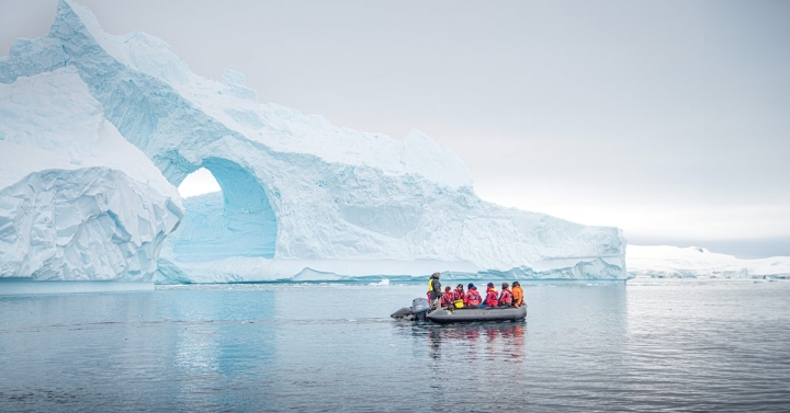 A boat filled with people in heavy red parkas floats by a glacier.