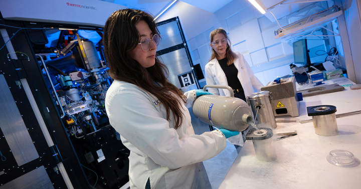 A scientist in a white lab coat and safety goggles pours liquid from one container into another. 