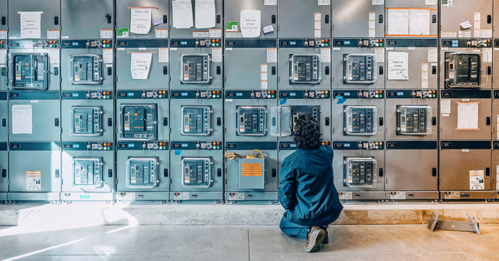 A scientist kneels in front of a wall of switchgear.