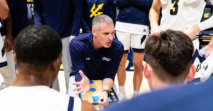 The men's basketball coach holding a clipboard and speaking to his team in a huddle.