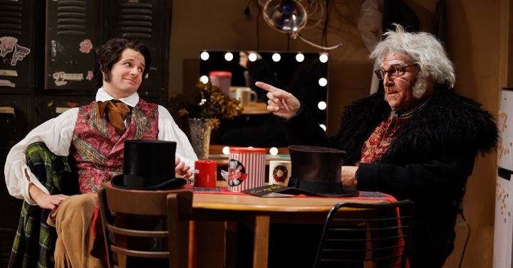 In Christmas Carol costumes and wigs, characters Eddie and Scrooge chat at table in theatre green room