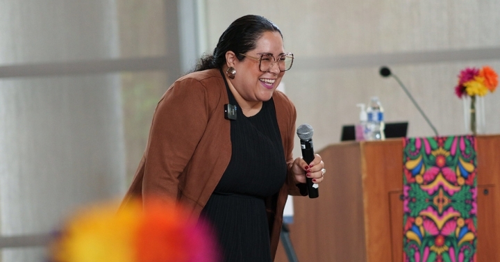 Dr. Dalia Pimentel laughs as she holds microphone and speaks to audience, with colorful flowers and tapestry pictured.