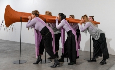 Six women blow into a large horn as part of a contemporary art exhibit.