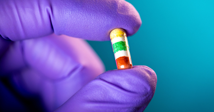 Small capsule with a yellow, white, green, white and red striped pattern held by a hand wearing a purple glove.