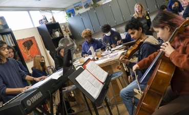 A Preuss sophomore plays keyboard while three other musicians join him on violin and viola as they play his original composition. Around them sit art students at wooden tables, painting their portraits as they play.