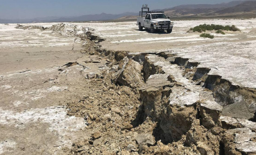 Surface rupture from the magnitude 7.1 Ridgecrest earthquake in July 2019. A giant crack is shown amid a dry desert landscape with a truck and mountains in the distance.