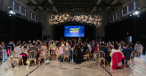 Video: People sit at round tables in a gym with a slideshow playing on a big screen at the front of the gym that says 