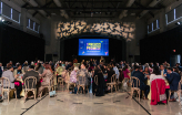Video: People sit at round tables in a gym with a slideshow playing on a big screen at the front of the gym that says 