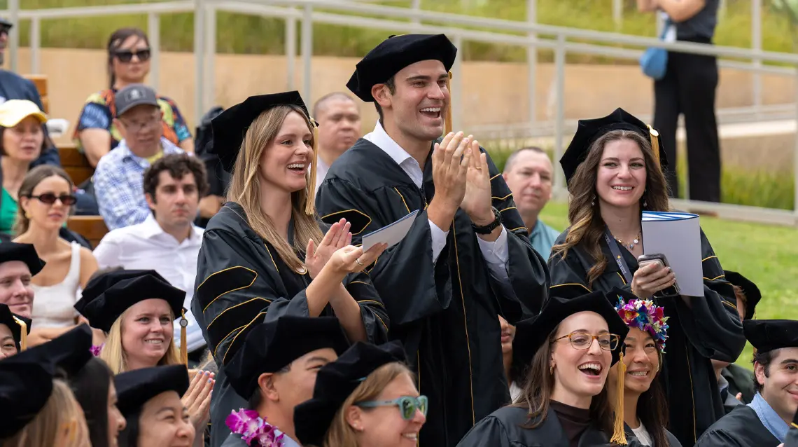 two female and one male student standing up clapping for their fellow graduate