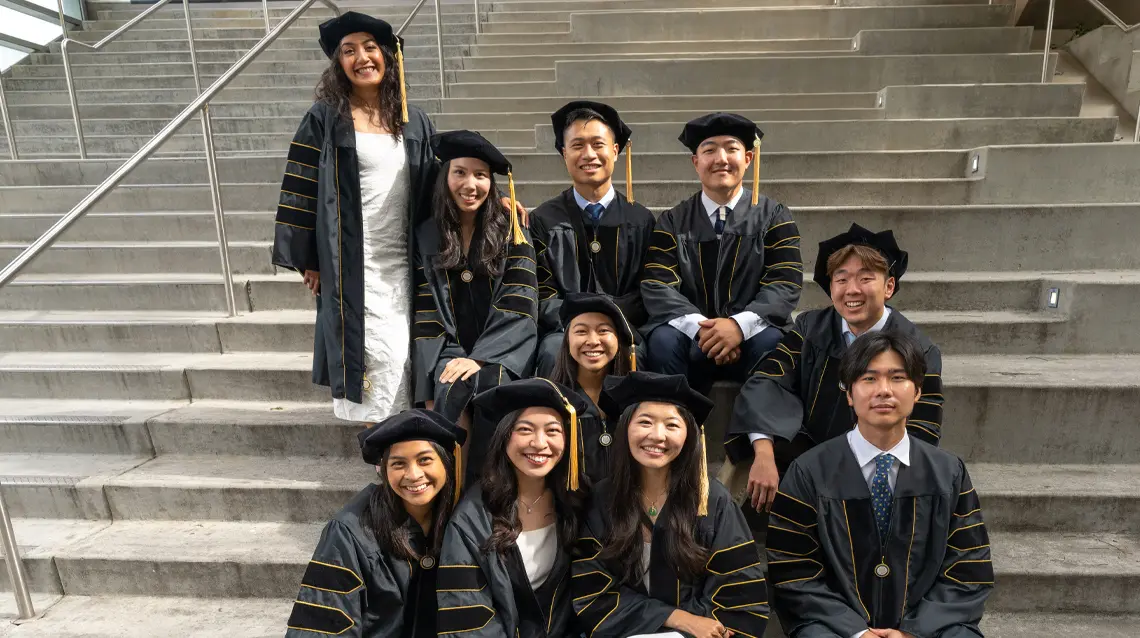 Group of male and female graduates sitting staggered on a concrete stairwell all smiling at camera