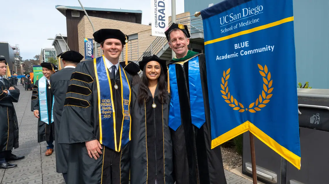 Paul Michael Acosta with a female classmate and male faculty member with the blue academic community sign.