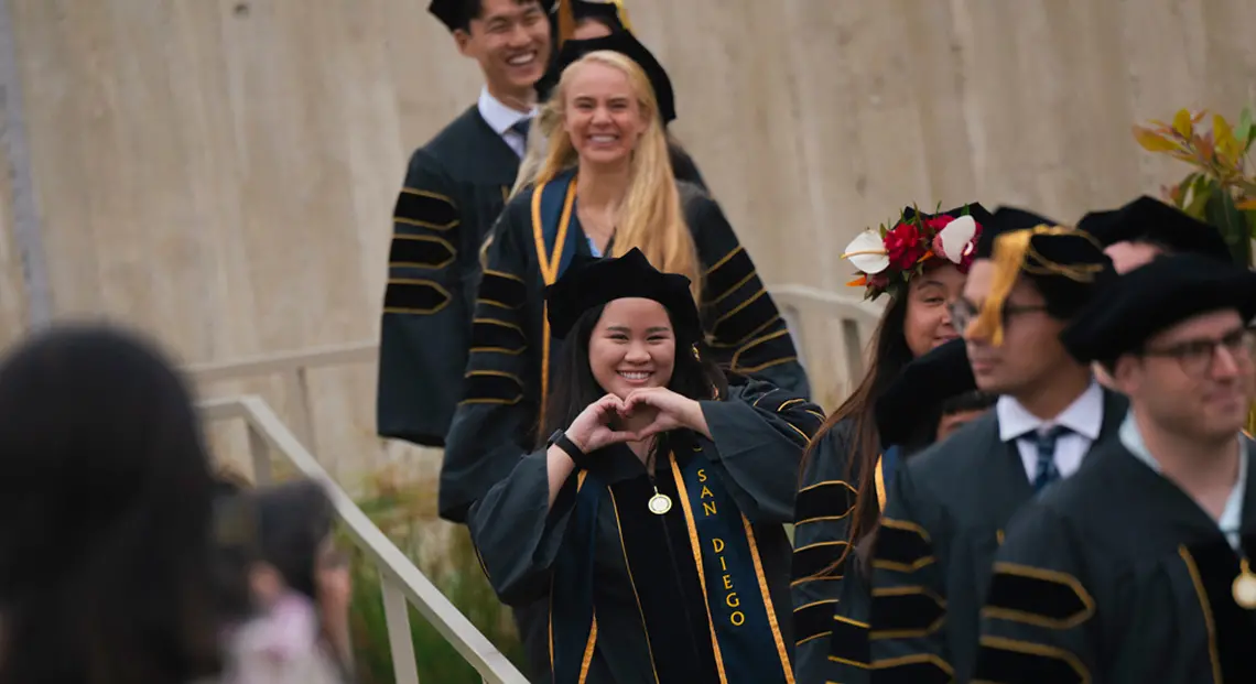 student walking into commencement ceremony female student forming heart with her hands smiling