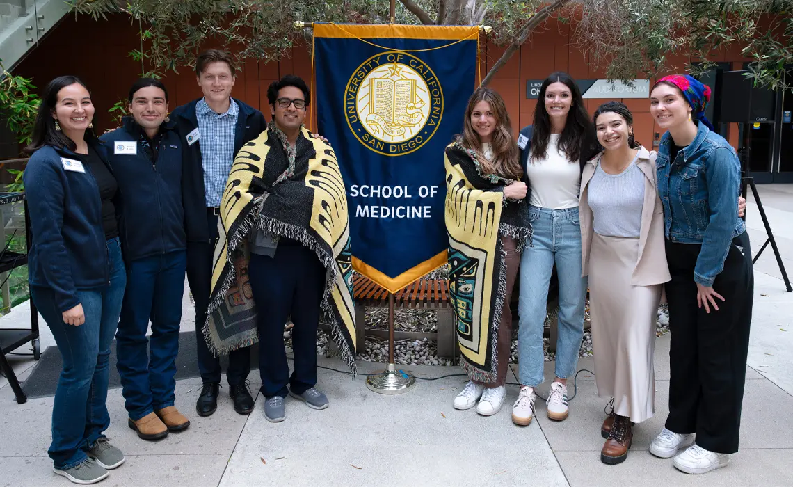 group photo of 8 medical students standing in a semi circle around a School of Medicine banner. They are all looking at the camera and smiling