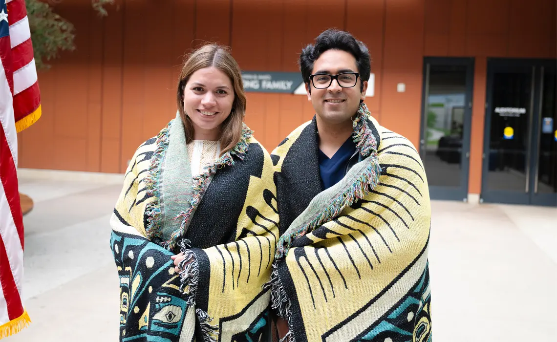 PRIME-TIDE students Gwendalynn Stilson and Cameron Salehi are shown standing next to each other smiling at the camera, each wrapped in their Native American blanket