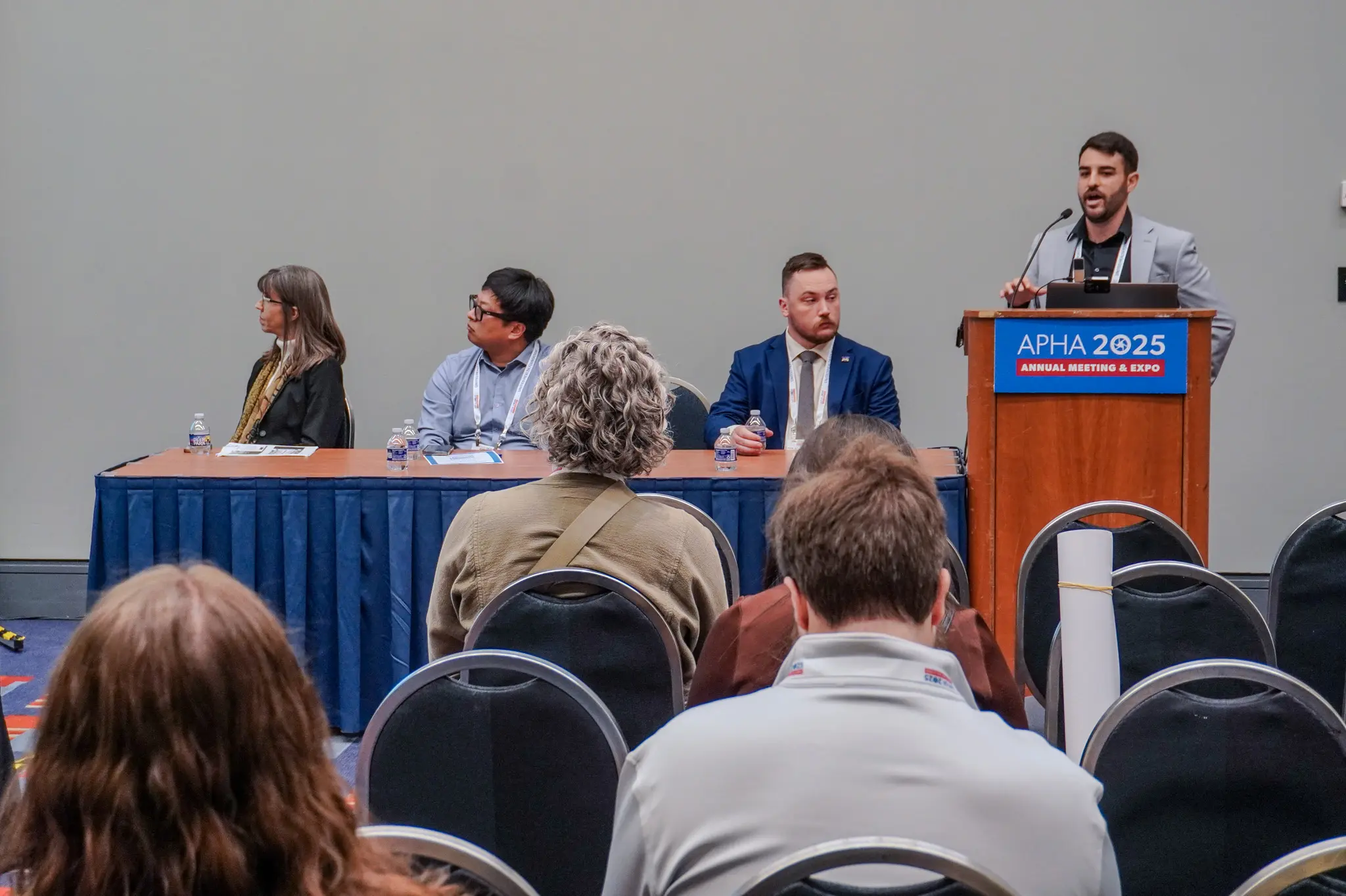 Jacob Carson stands at a podium speaking while other panelist sit at a table