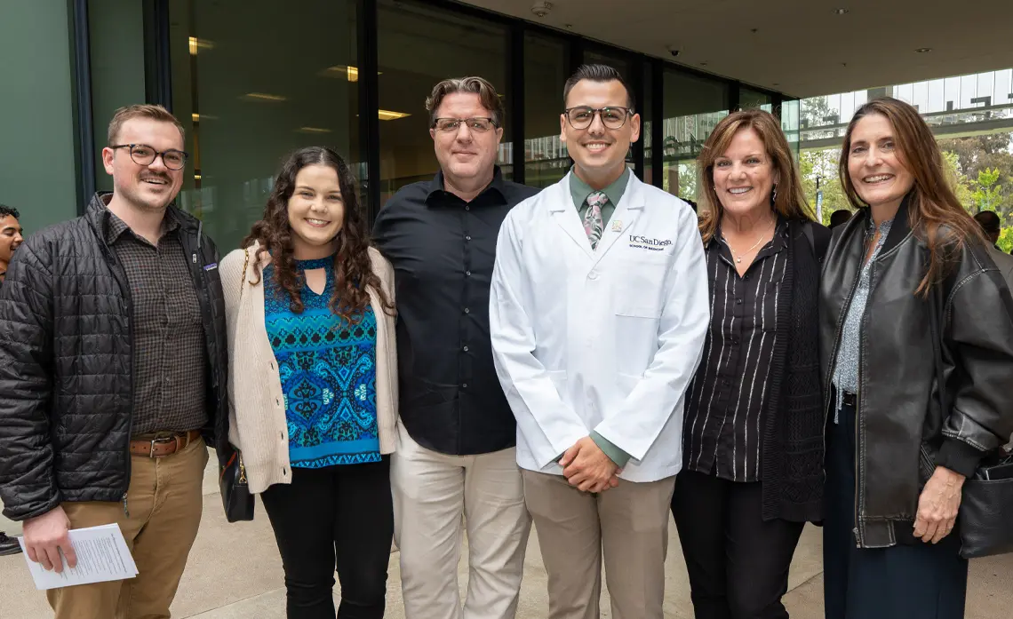 Male student wearing white coat surrounded by family and friends all smiling and looking at the camera.