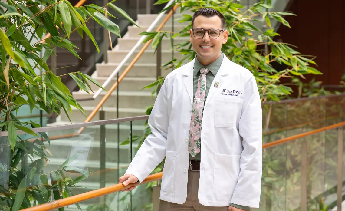 Male student wearing a white coat standing by a tree posing and smiling towards the camera