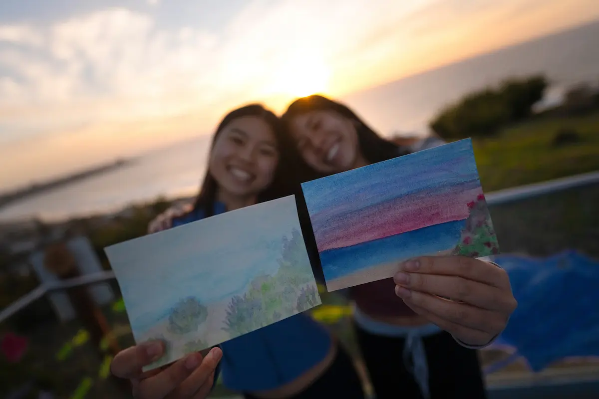 Two students hold watercolor paintings up to camera