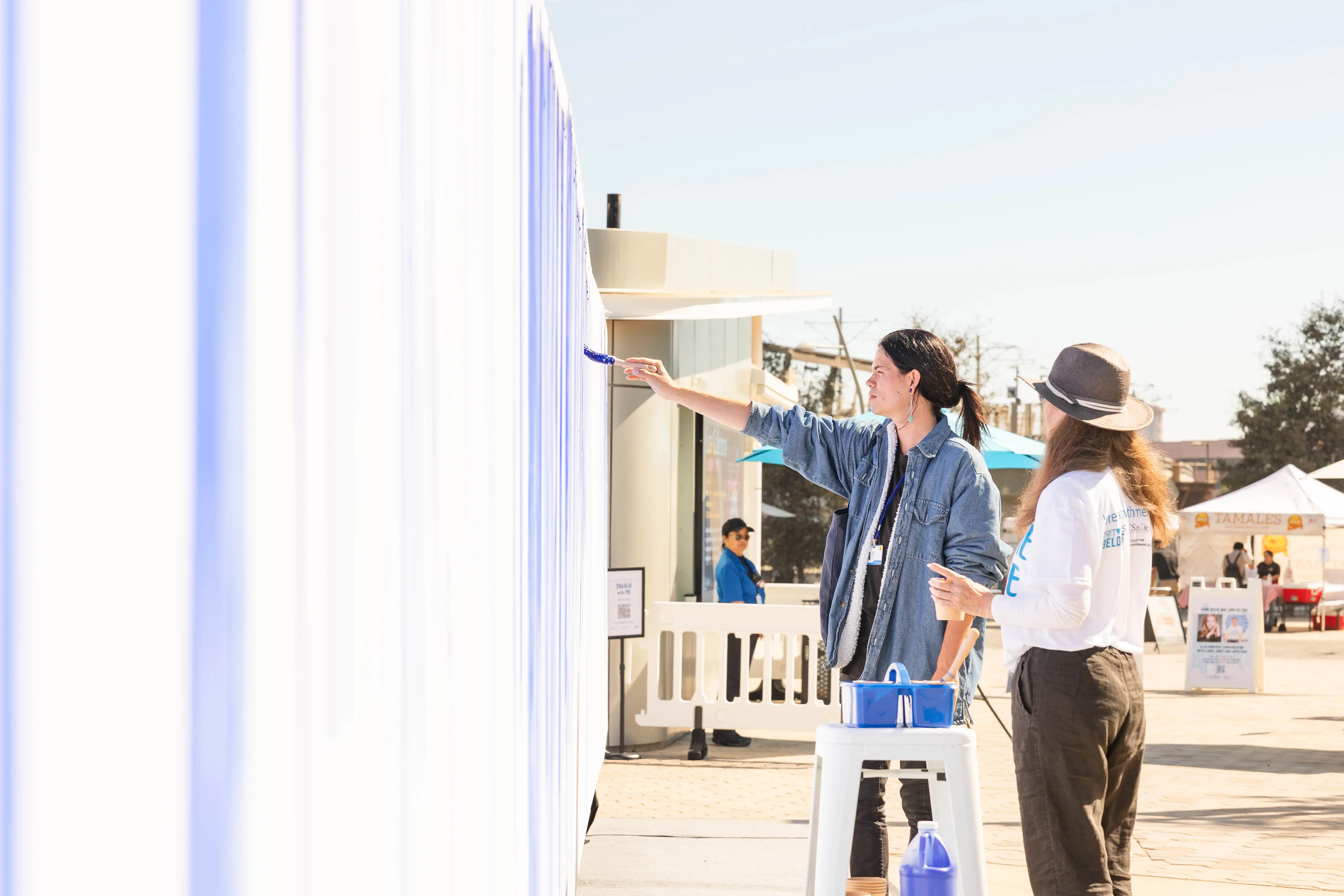 A person paints a line on the canvas as a volunteer looks on.