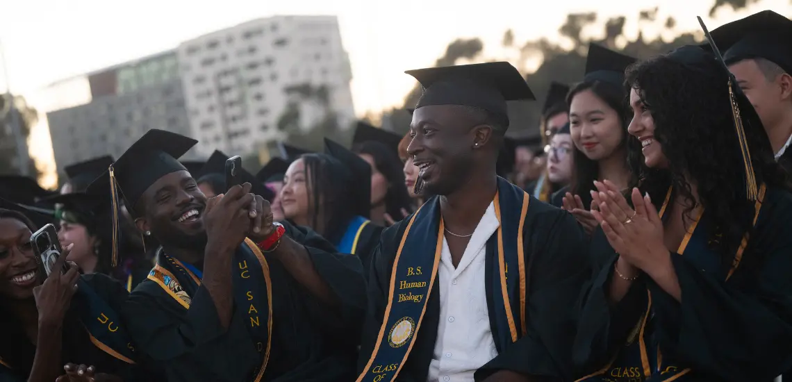 Student in cap smiles while another graduate takes picture with phone