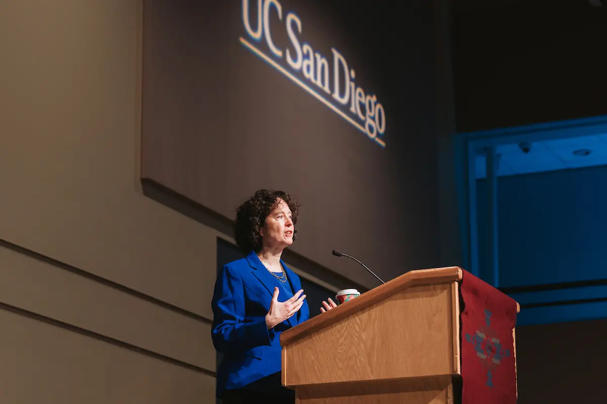 A woman with short brown hair and a blue blazer speaks at a podium