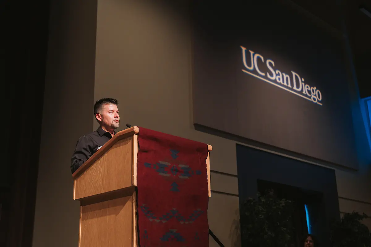 A man with black hair wearing a black shirt speaks from behind a podium.