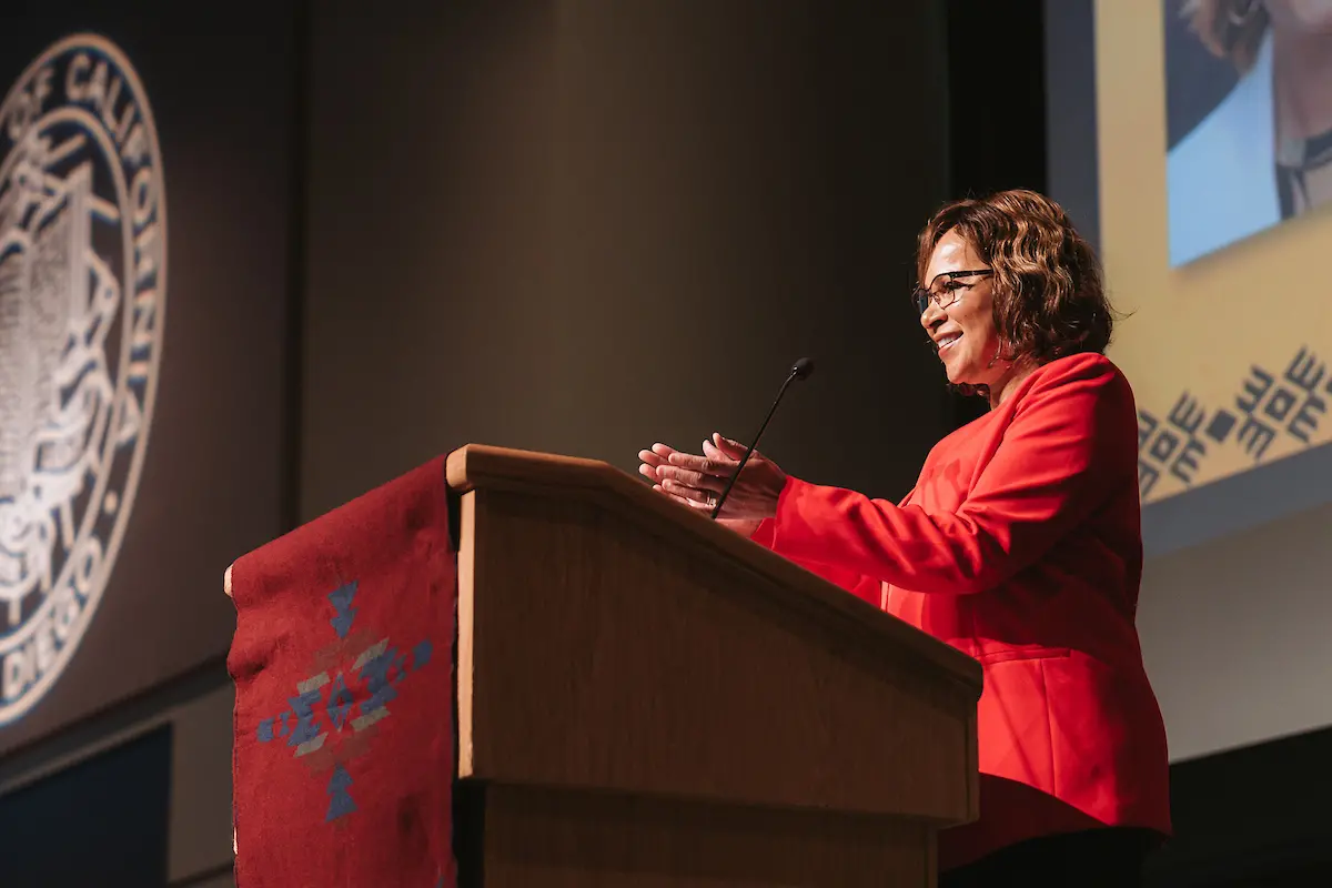 A woman with brown hair wearing a red blazer claps and smiles at the crowd from behind a podium.