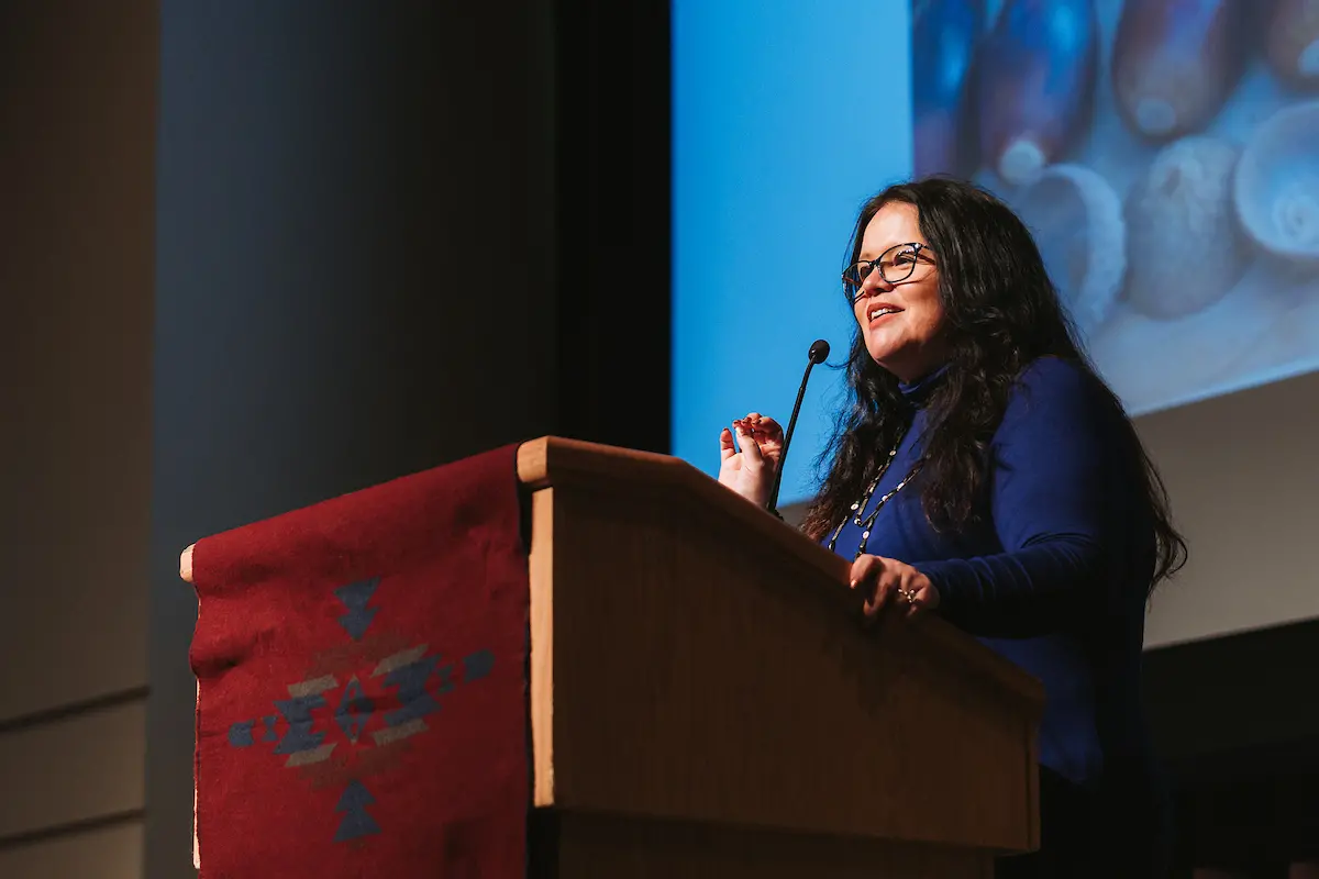 A woman with black hair wearing a blue shirt speaks from behind a podium.