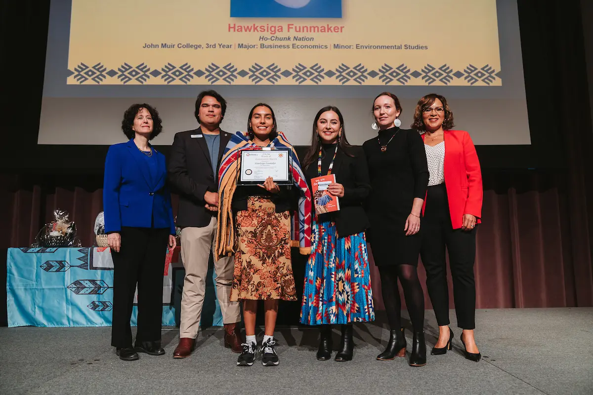 A person with a blanket wrapped around them holds a certificate and stands on stage next to five others.