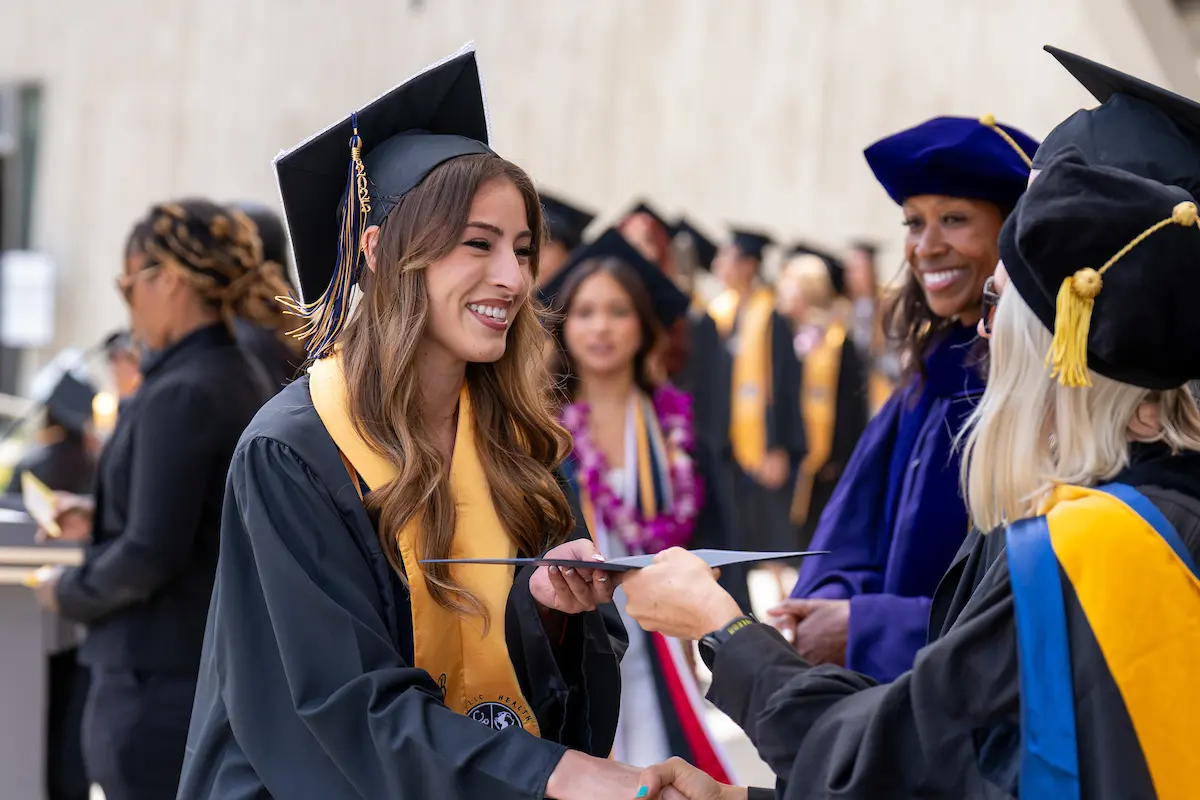 Image is of a Bachelor of Science in Public Health Student receiving their diploma. They are wearing their graduation regalia and a gold sash that reads: