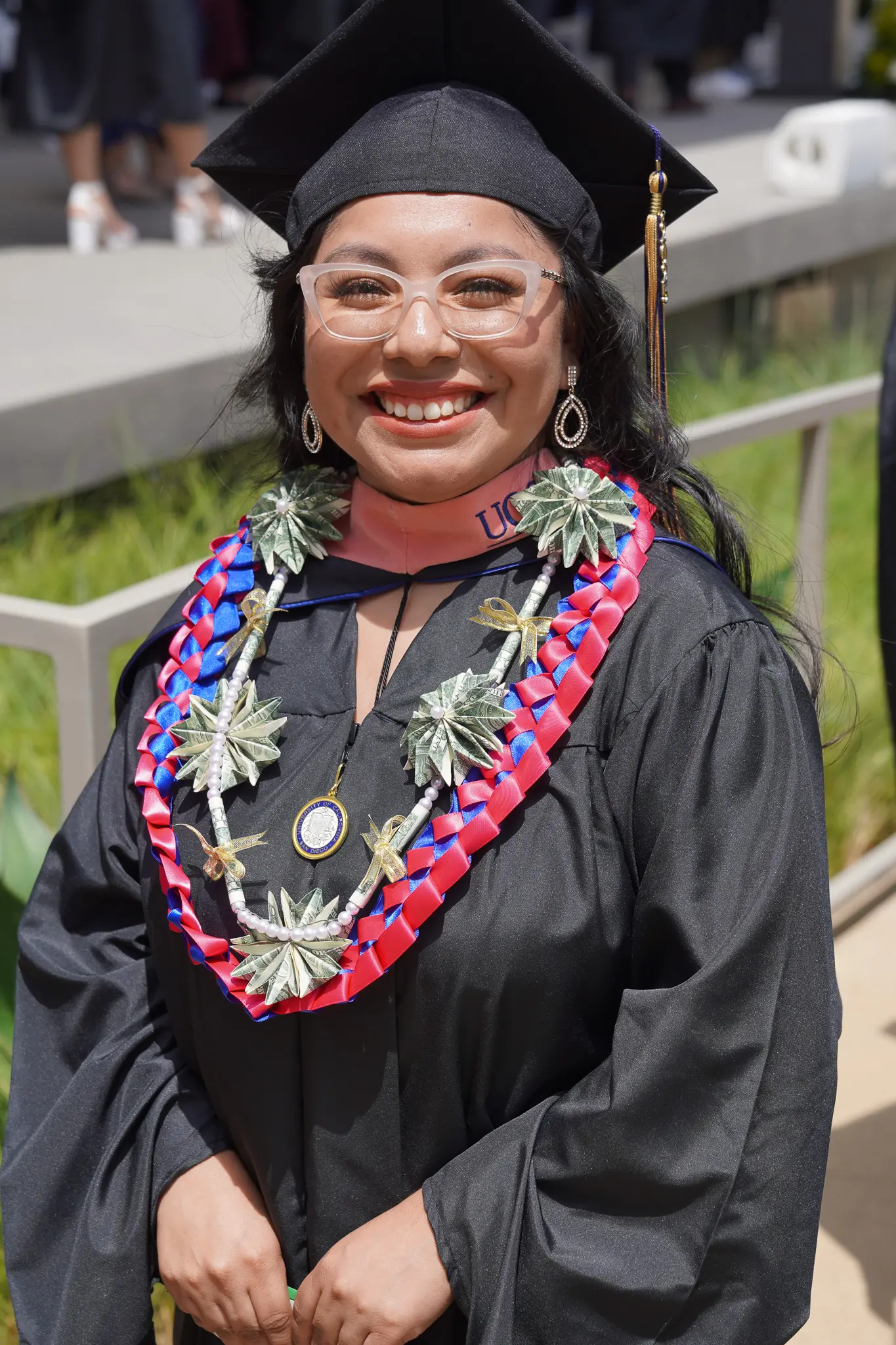 Photo of a graduate as they are walking back to their seat. They are wearing traditional black and gold graduation regalia. They are wearing glasses, and a lei.