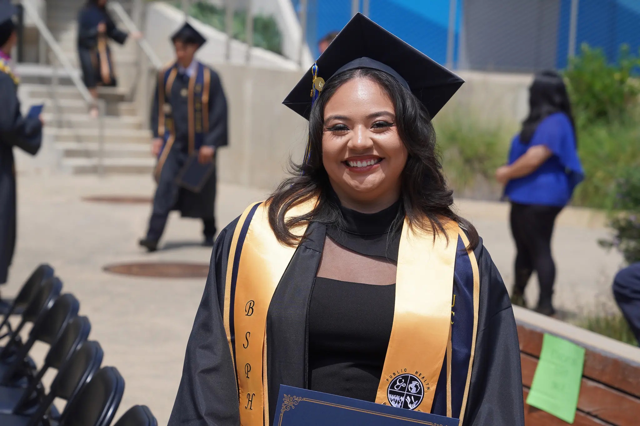 Photo of a graduate as they are walking back to their seat. They are wearing traditional black and gold graduation regalia. There is a golden sash that reads: