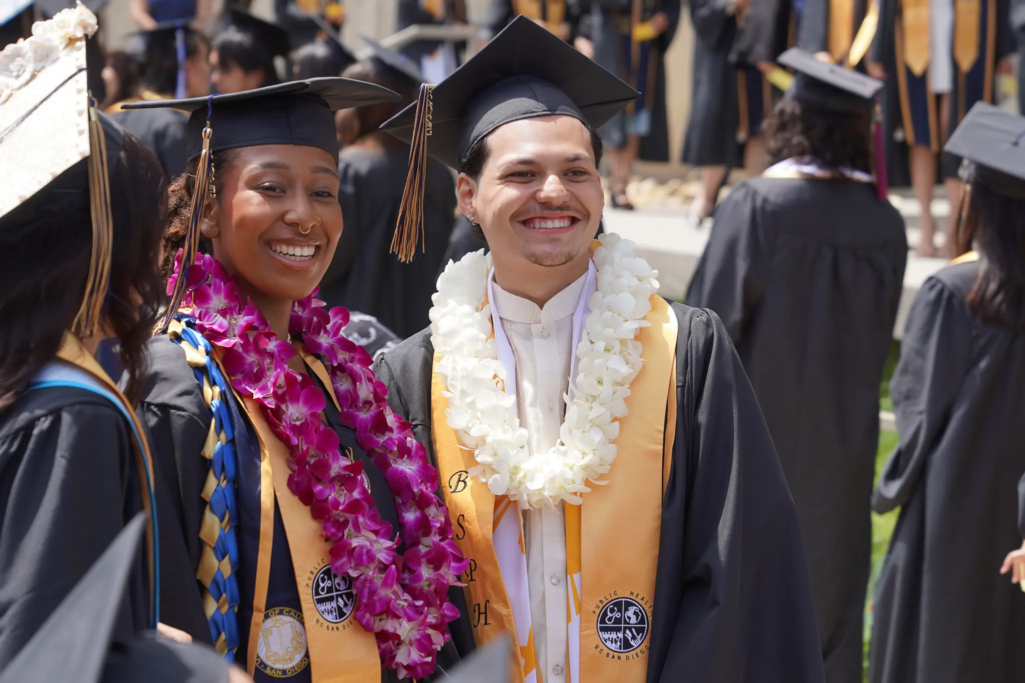 Two Bachelor of Science in Public Health students are standing shoulder-to-shoulder in their graduation regalia. Their sashes read: