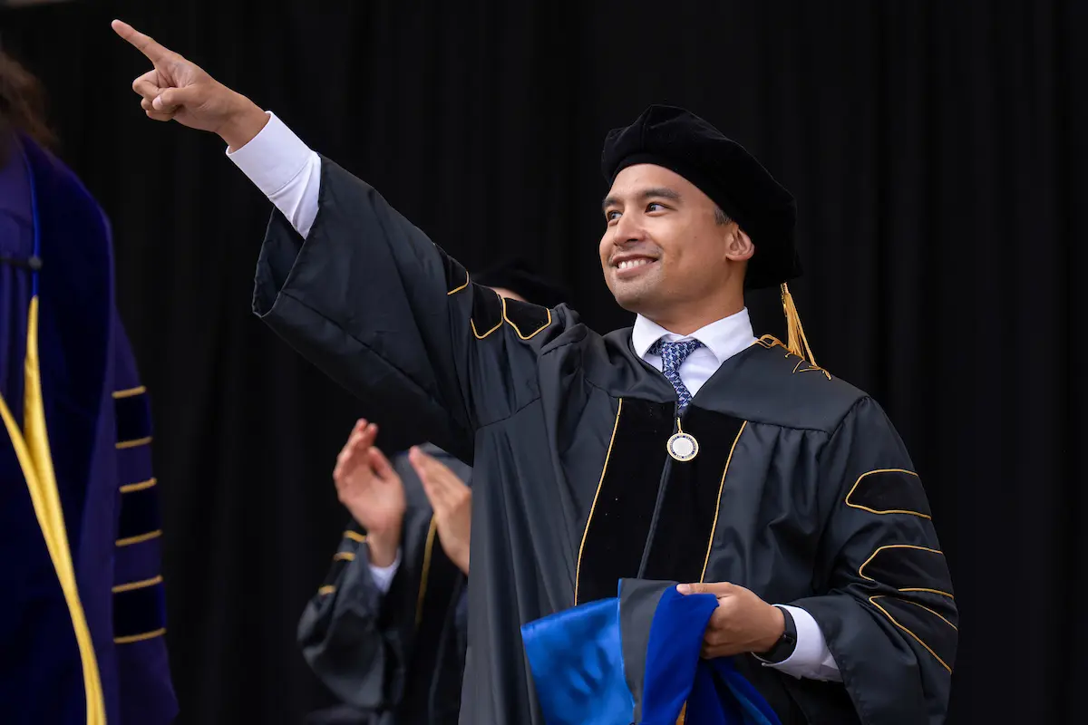 Doctoral student is pointing up to their family in the stands. They are wearing traditional black graduation regalia and holding a blue hood.