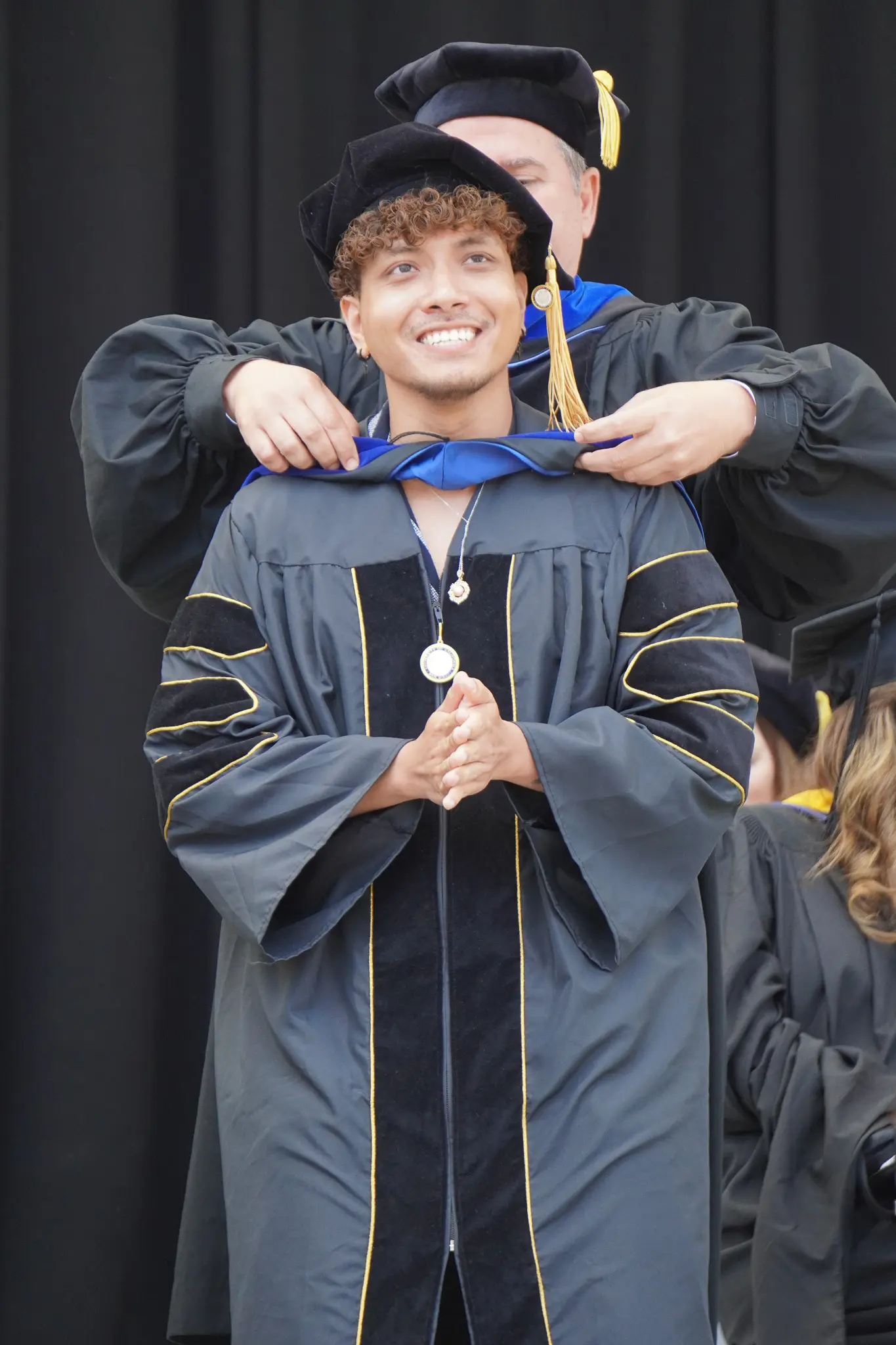 Doctoral student who receiving their hood. Their hands are clasped together. They are wearing traditional black graduation regalia.