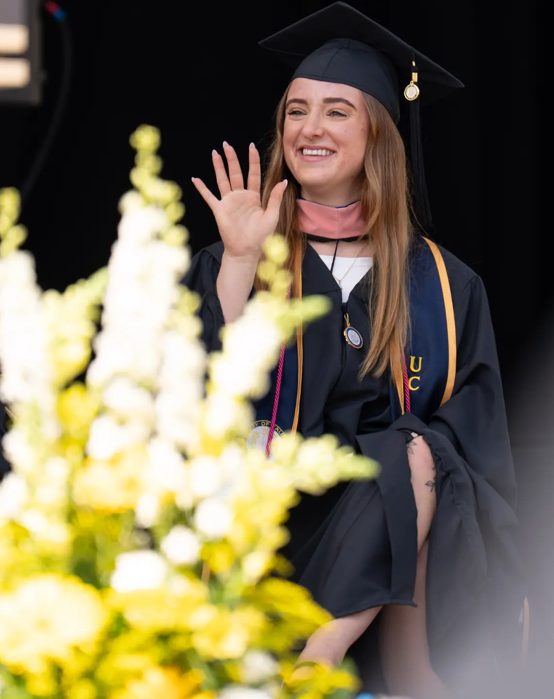 Naomi Rose Eisner Guy on stage. There are yellow and white flower in the foreground. Naomi is waving, wearing traditional graduation regalia.