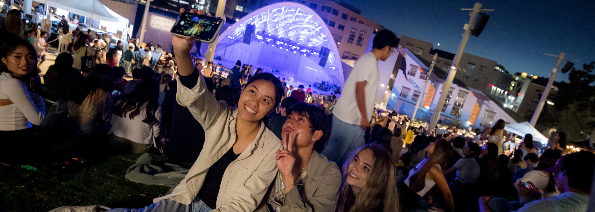 A group of three students sit on the grass at the Epstein Family Amphitheater outdoors at night while posing for a selfie image with their cell phone.