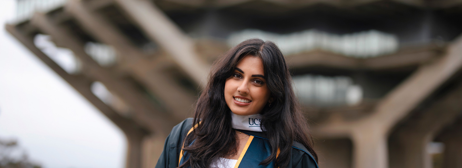 A smiling scholar in commencement regalia standing in front of a library