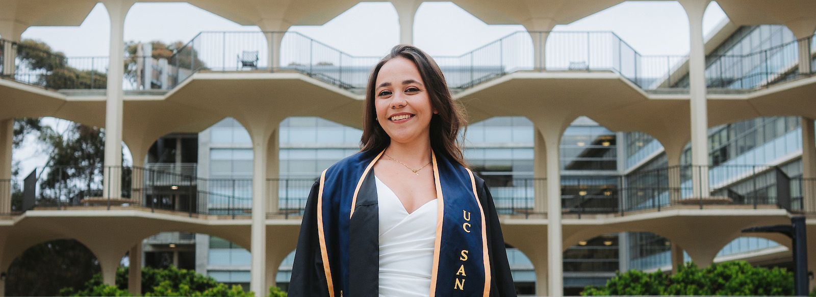 A student in commencement regalia smiling in front of a unique building