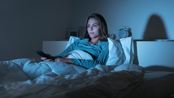 Woman sits in bed with remote in hand, face illuminated by light of TV