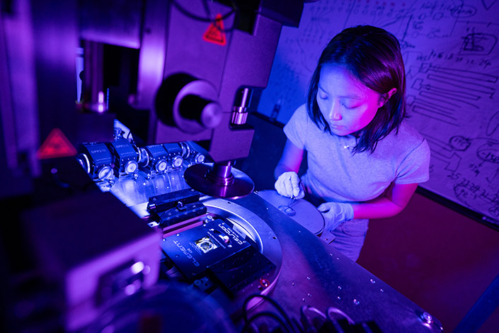 Woman looking down and pointing with tweezers at a small round metallic disk. A microscope is in the foreground.