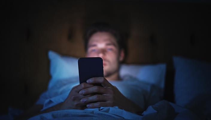 Man's face illuminated by glow of smartphone as he lounges in bed in dark