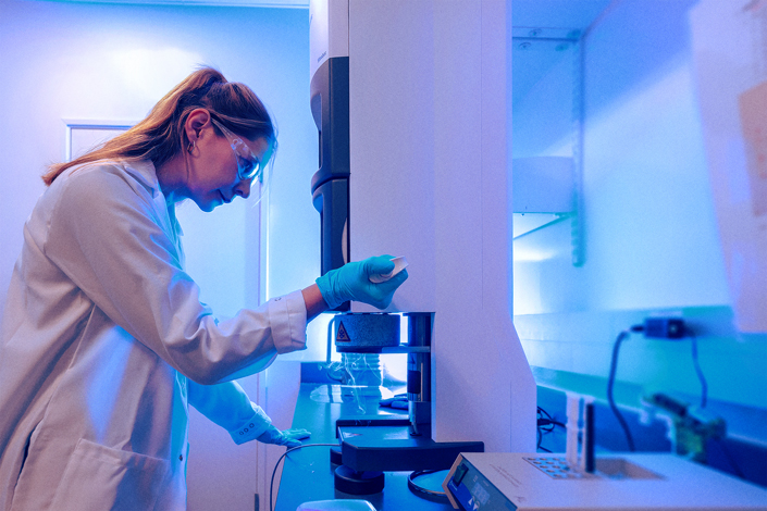 A scientist in a white lab coat uses a pipette to prepare a sample while standing in front of a large piece of equipment.