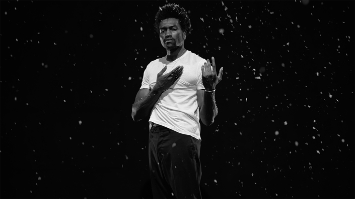 A man in a white t-shirt and black pants gestures while standing against a black background.