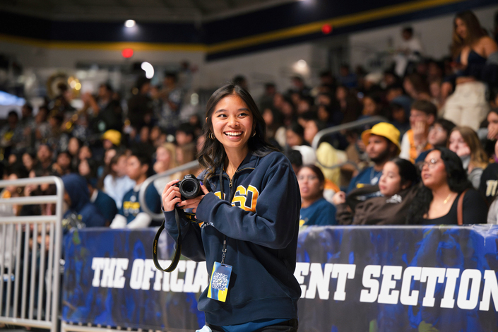 A woman in a sweatshirt holding a camera stands courtside in front of tiered audience seating.
