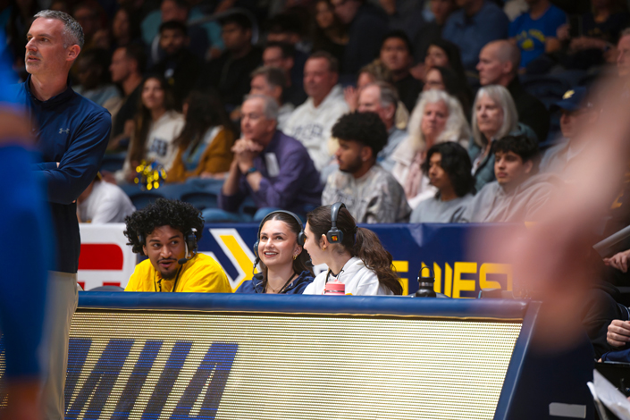 Three people sit at a courtside table in front of tiered seating.