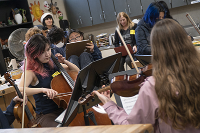 Four musicians are pictured, one playing cello, two violin and one viola. Behind them student artists paint their portraits.